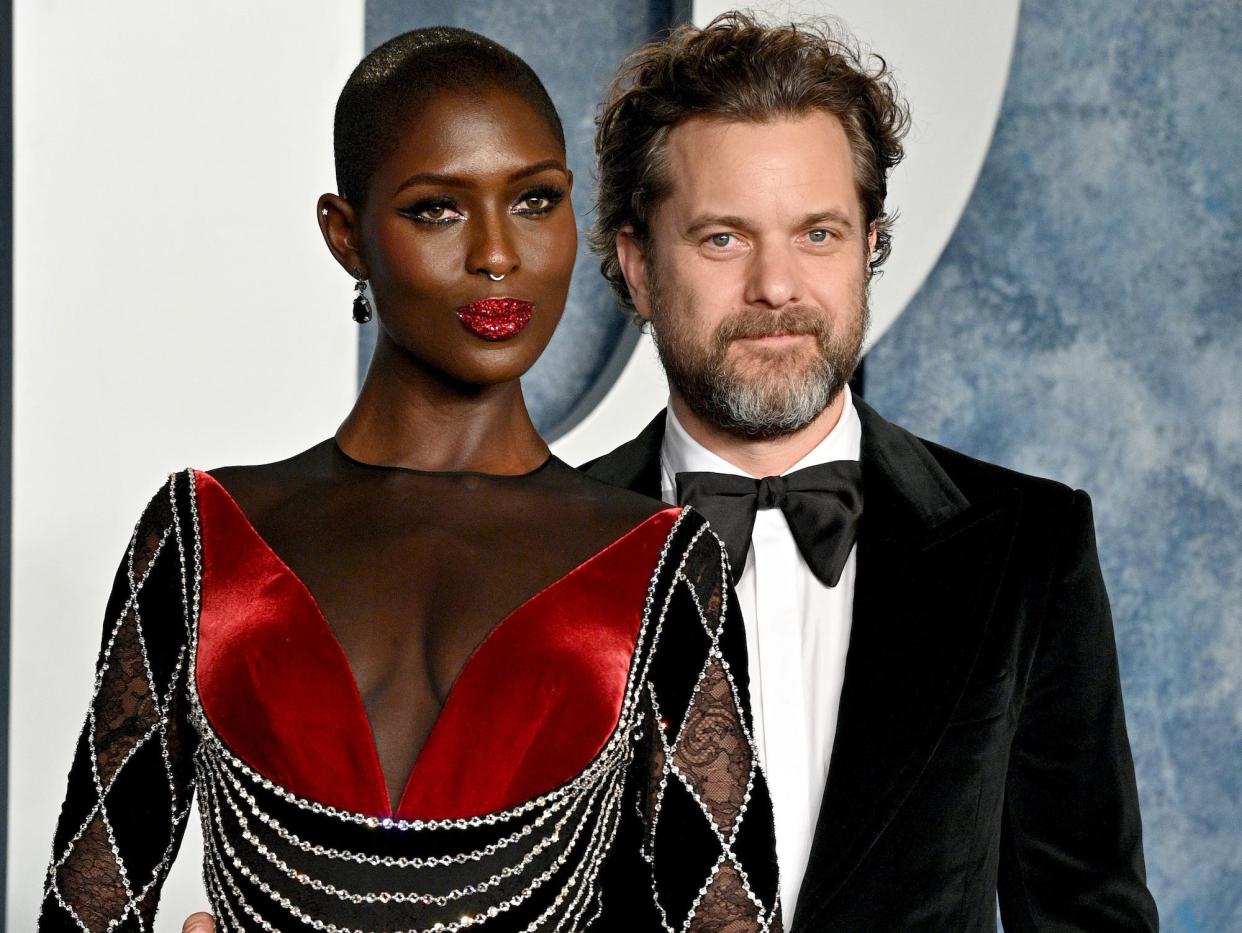 Jodie Turner-Smith, in a black and red sheer gown with rhinestone accents and matching red lipstick, poses on the red carpet with husband Joshua Jackson, who is wearing a black velvet tux and has his arm around Turner-Smith's waist.