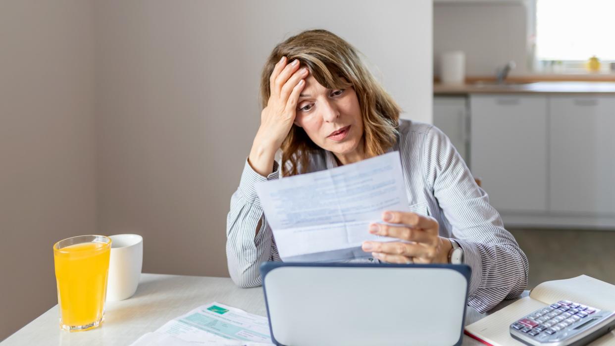 Stressed and Worried Senior Woman Calculating Domestic Expenses, Sitting at Dining Table in Front of Open Laptop Computer.