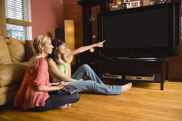 Two women sit on the floor of a living room, while one points to a large TV screen.