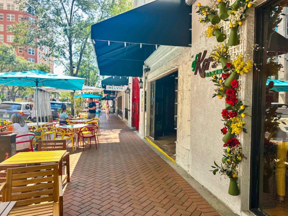 A walkway with brown bricks and tables and chairs next to shops with a flower display in the foreground