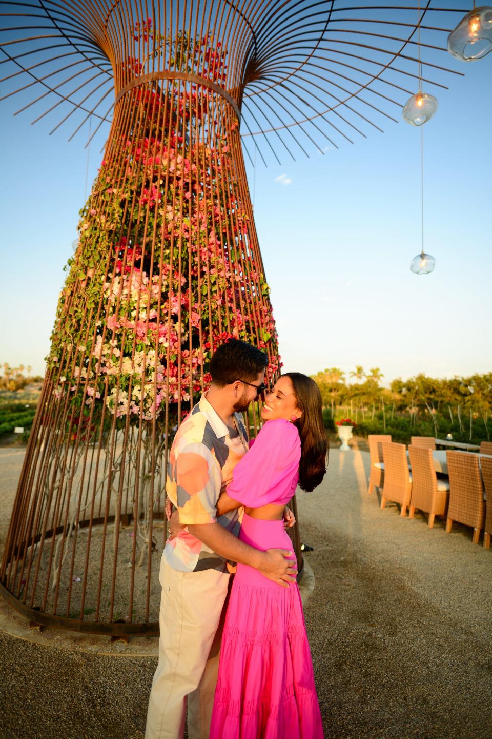 A man and woman embrace in front of a floral sculpture.