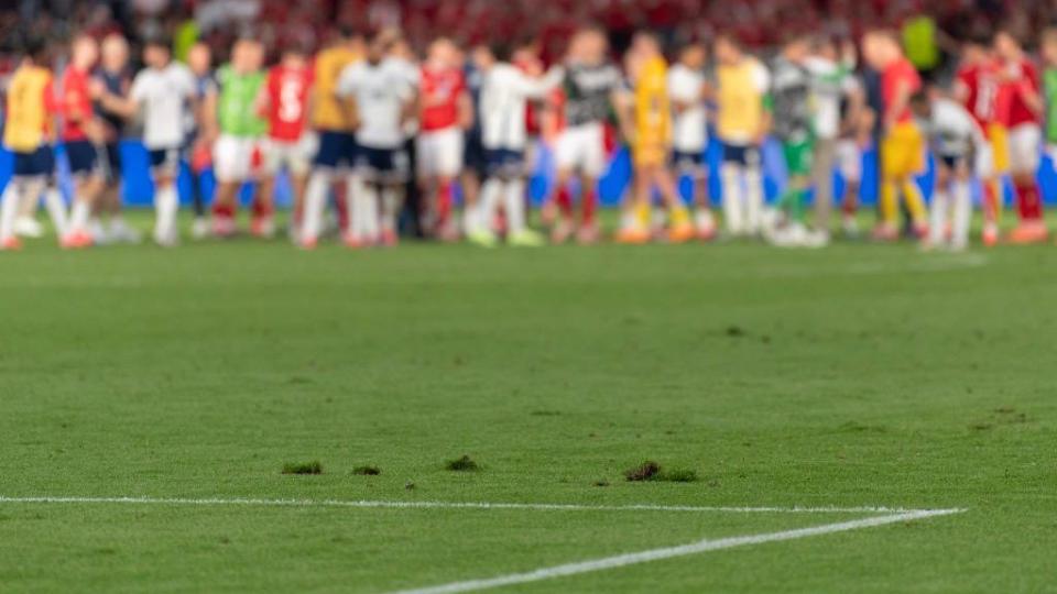 Lumps of grass on top of the pitch with players in the background