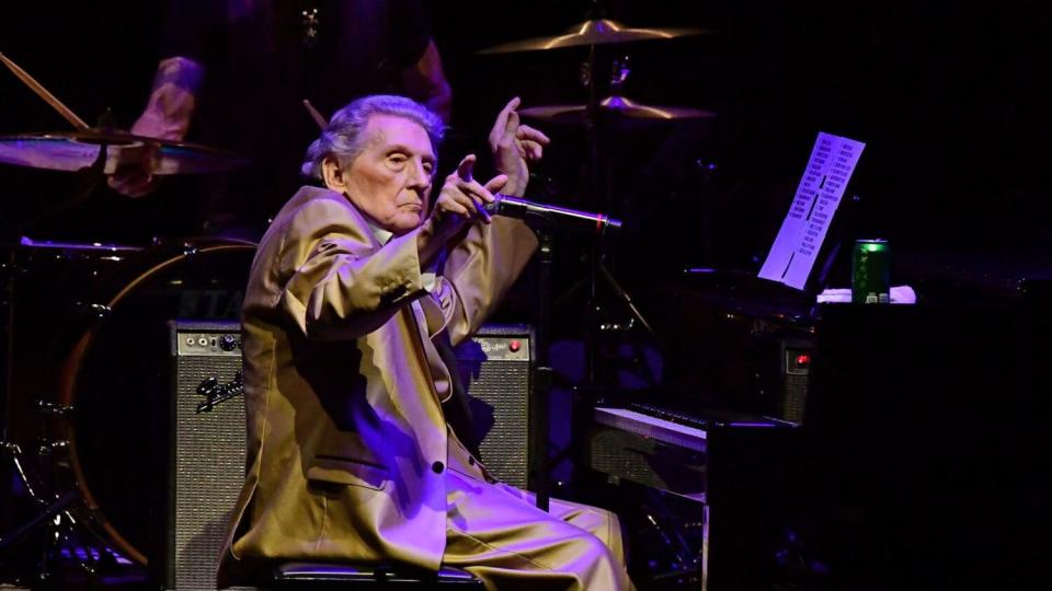 Jerry Lee Lewis, in a gold suit, gestures while seated at the piano on stage.