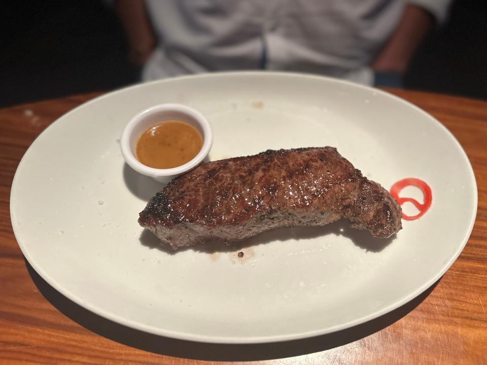 A New York strip steak on a large white plate with a red sauce and a small white cup of brown sauce on the side