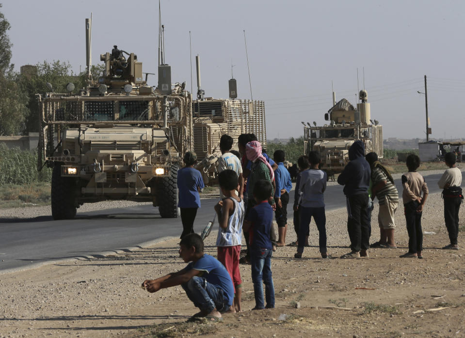 FILE - In this July 26, 2017, file photo, Syrians look at a U.S. armored vehicle convoy on a road that leads to Raqqa, northeast Syria. With Islamic State's near total defeat on the battle field, the extremist group has reverted to what it was before its spectacular series of conquests in 2014 _ a shadowy terror network that targets vulnerable civilian populations and exploits state weaknesses to incite on sectarian strife. But a recent surge in false claims of responsibility for attacks also signals that the group is struggling to stay relevant after losing its proto-state and its dominance of the international news agenda. (AP Photo/Hussein Malla, File)