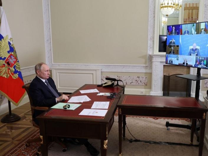 Russian President Vladimir Putin speaking to a raised TV with the images of Russian government officials while sitting behind a large wooden desk