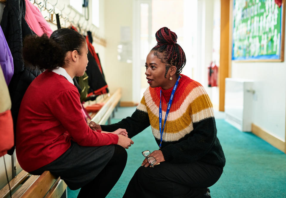 Teacher kneels to speak with a young student in a school hallway, both are engaged in conversation