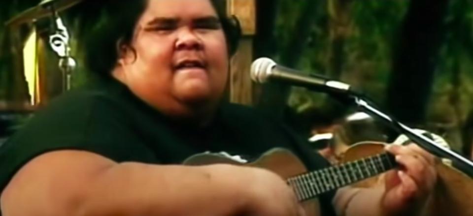 Israel Kamakawiwo'ole playing a ukelele and singing to a mic.