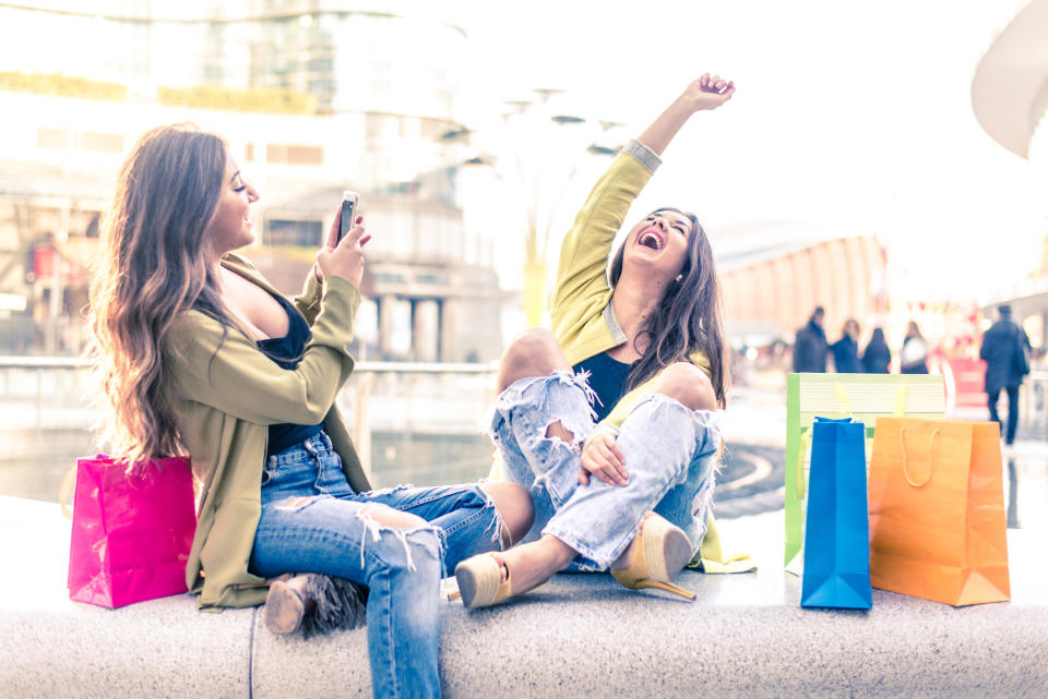 Two teen girls sitting with shopping bags and laughing while one is taking pictures of the other.