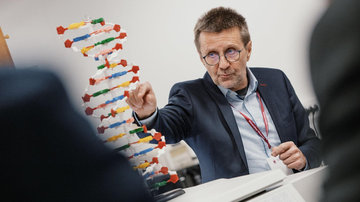  Virginijus Šikšnys (an older man with short brown hair and round glasses wearing a suit jacket) gestures toward a 3D model of DNA on a table. 