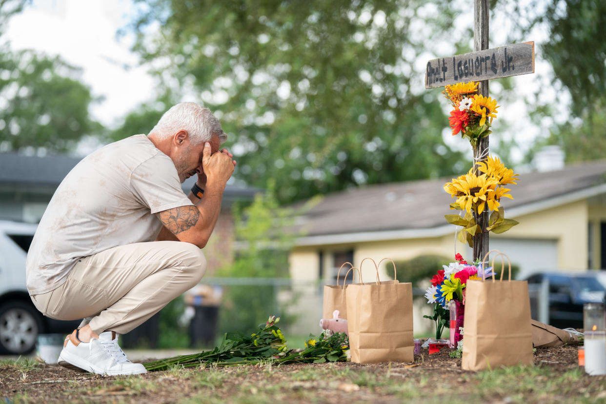 Will Walsh squats next to a wooden cross decorated with flowers with his head in his hands.