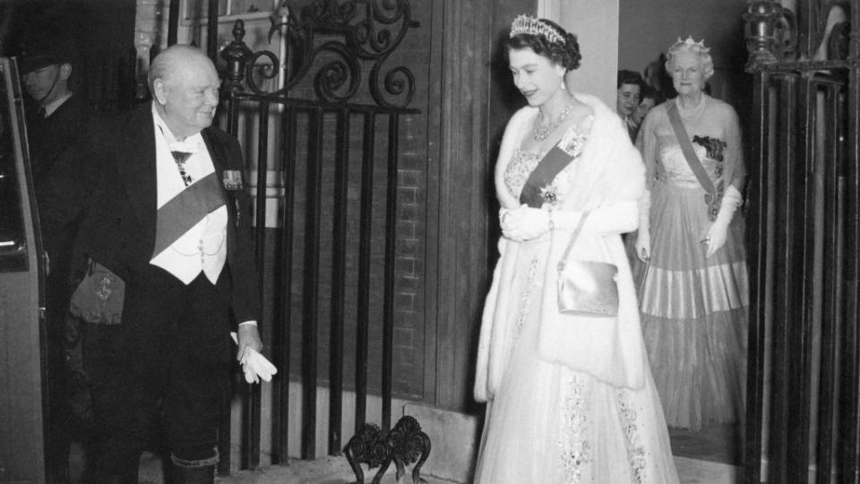 winston churchill holds a car door open and watches queen elizabeth walk toward it, he wears a tuxedo with a sash, she wears a gown with a fur stole, sash, and crown