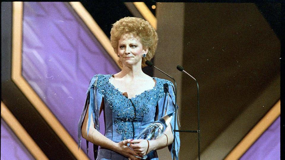 reba mcintire crying while standing on a stage, she holds a crystal award in her arms and stands behind a microphone on a stand, she wears a blue dress