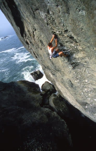 Man climbs steep gray stone above a crashing ocean.