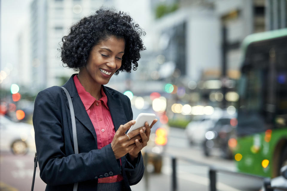 Smiling young businesswoman surfing net on mobile phone. Female commuter is standing in financial district. She is having black curly hair.