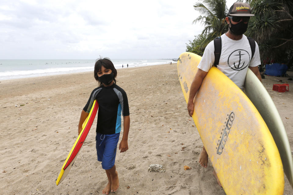 Tourists wearing face masks carry their surfboard at Kuta beach, Bali, Indonesia on Thursday, July 9, 2020. Indonesia's resort island of Bali reopened after a three-month virus lockdown Thursday, allowing local people and stranded foreign tourists to resume public activities before foreign arrivals resume in September.(AP Photo/Firdia Lisnawati)