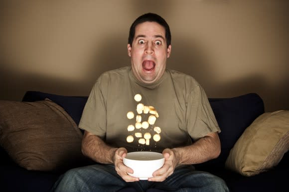 A man on a sofa sits with an open-mouthed expression of surprise, as the food in his bowl flies into the air.