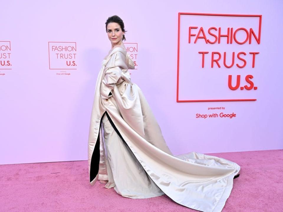 sophia bush in a flowing, voluminous white gold gown, her hair worn in an updo, and looking back over her shoulder on a pink carpet