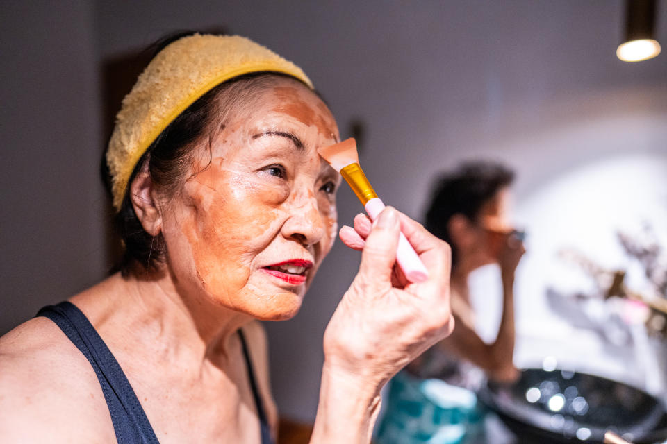 Elderly person applying makeup with a brush, standing in front of a mirror, embodying self-care