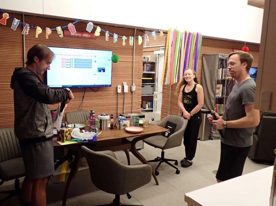 three chapea crew members stand around a table with snacks below a happy birthday banner while one opens a package