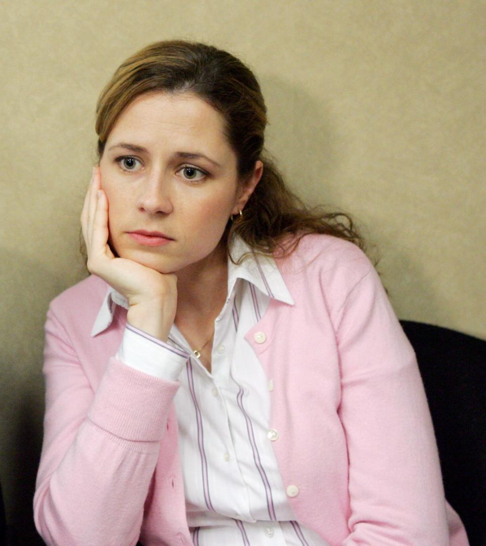 Pam from The Office sitting against a wall, looking forward with mild expression