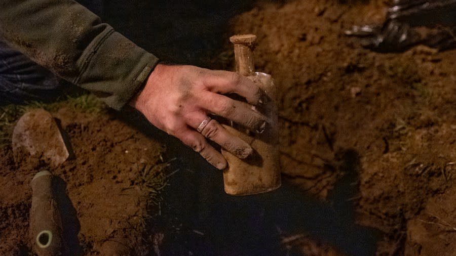 A man pulls a small glass bottle filled with dirt and seeds out of a hole.