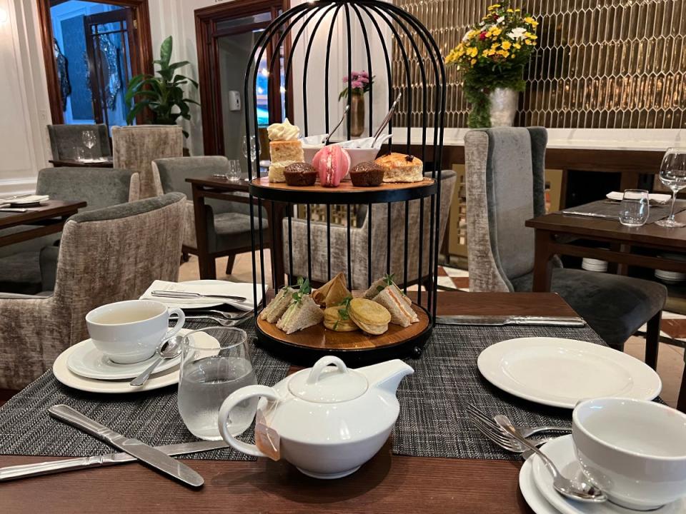 A table with tea cups and tea pot with a tower of pastries and sweets on the table.  Wooden tables with gray chairs and a metallic wall are in the background