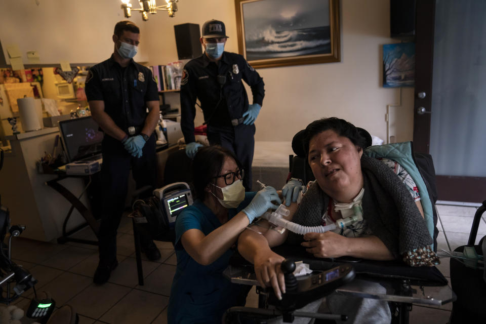 Pharmacist Stella Kim, foreground left, administers the second dose of the Pfizer COVID-19 vaccine to Socorro Franco-Martinez, who has muscular dystrophy, in her apartment as Torrance firefighters, Trevor Borello, rear left, and Alessandro Demuro watch Wednesday, May 12, 2021, in Torrance, Calif. Teamed up with the Torrance Fire Department, Torrance Memorial Medical Center started inoculating people at home in March, identifying people through a city hotline, county health department, senior centers and doctor's offices, said Mei Tsai, the pharmacist who coordinates the program. (AP Photo/Jae C. Hong)