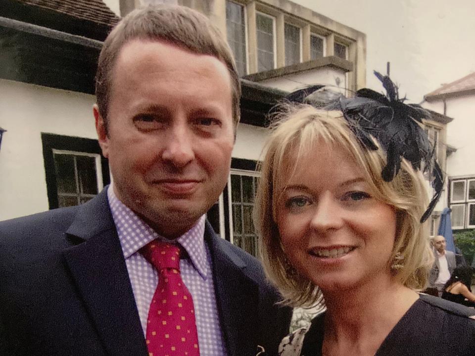 A man and woman at a wedding. The man is wearing a suit jacket. The woman is wearing a black dress and a fascinator.