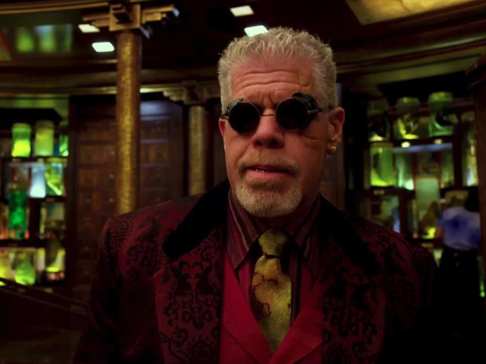 ron perlman as hannibal chau in pacific rim. he's a middle aged man wearing a deep red suit jacket, red vest, golden tie, and sunglasses with side filters. he's standing in a high tech lab filled with glowing green vials and specimens