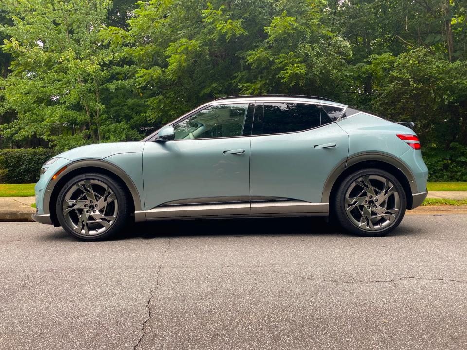 The side profile of a green 2024 Genesis GV60 EV parked in front of a row of trees.