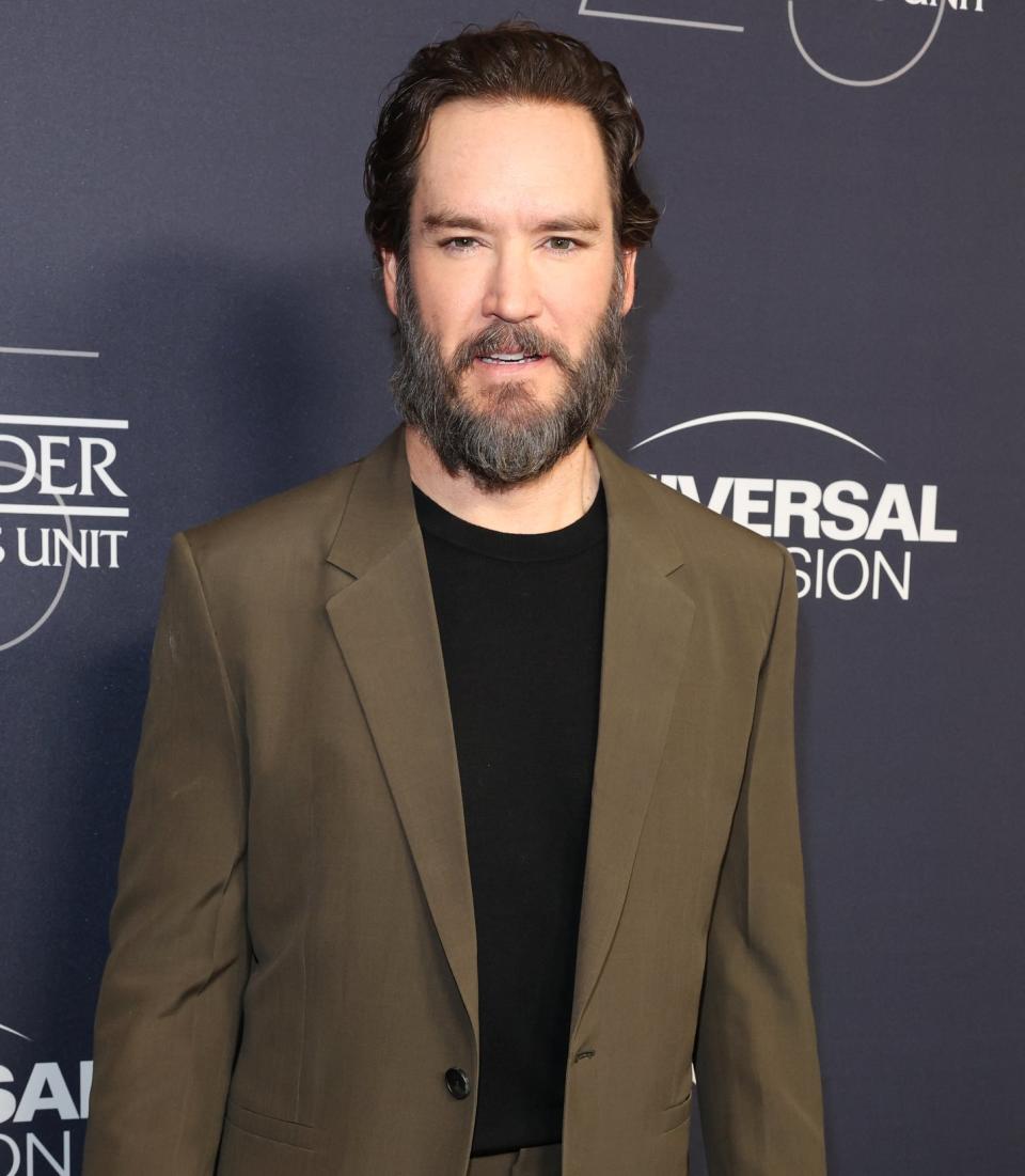 Mark-Paul Gosselaar at an event, wearing a dark suit over a black shirt, standing in front of a backdrop with logos including NBC and Universal Television