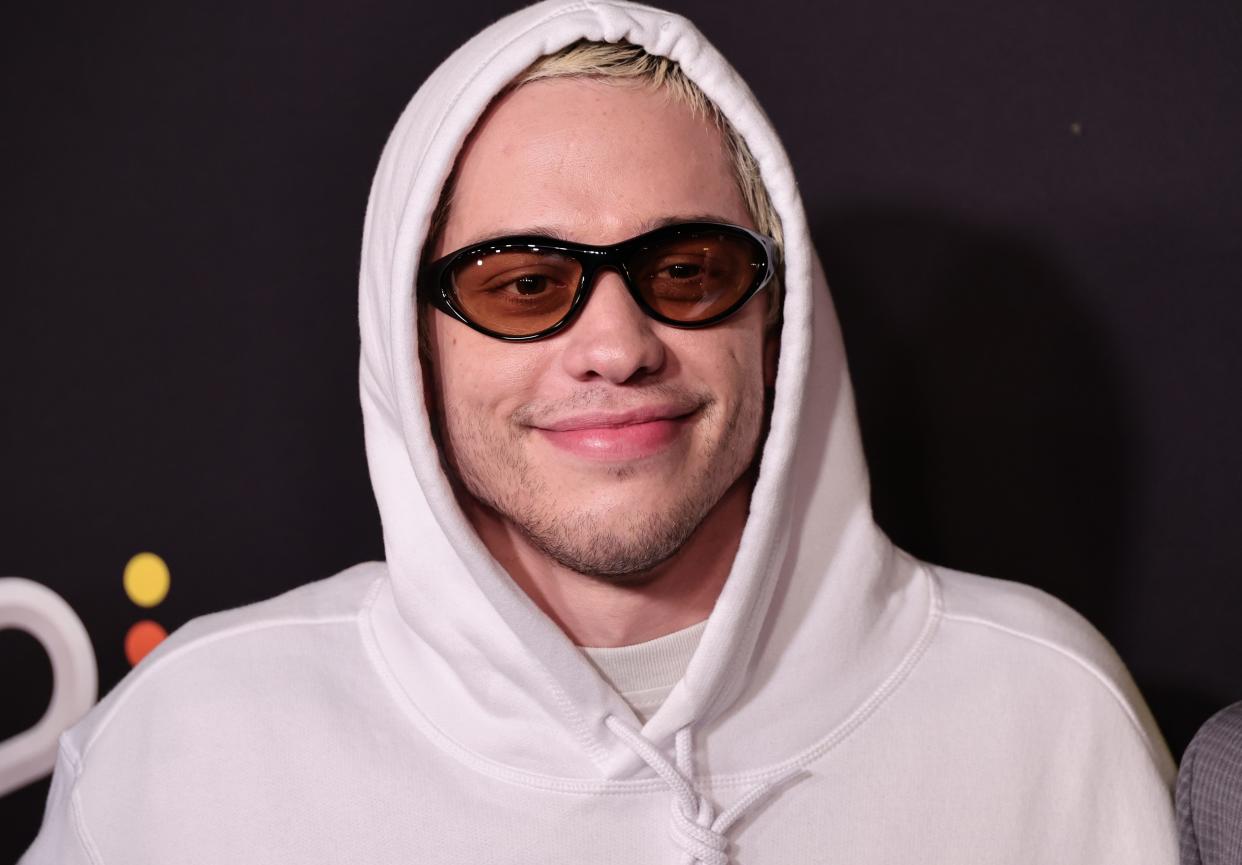 pete davidson wearing yellow tinted sunglasses and a white hoodie, pulled over his head and smiling