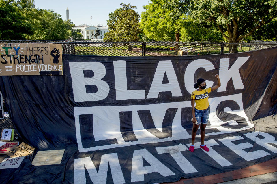 FILE - In this June 8, 2020, file photo, the White House is visible behind a woman who holds her fist up as she poses for a photograph with a large banner that reads Black Lives Matter hanging on a security fence in Washington, after days of protests over the death of George Floyd. (AP Photo/Andrew Harnik, File)