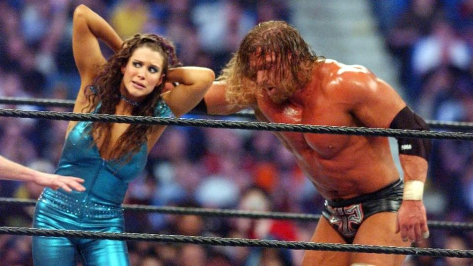 Wrestler Triple H grabs Stephanie McMahon by the hair in the middle of a wrestling ring. They both have light-toned skin and are in front of a large crowd.