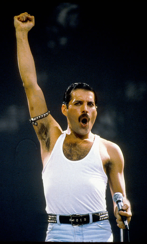 Freddie Mercury performs on stage, wearing a white tank top and white belted pants, raising his fist passionately while holding a microphone