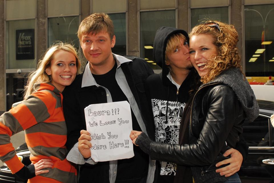 The brothers Nick and Aaron Carter stand between two smiling women, holding up a small paper sign that reads in handwritten marker "laura! we love you. wish you were here."