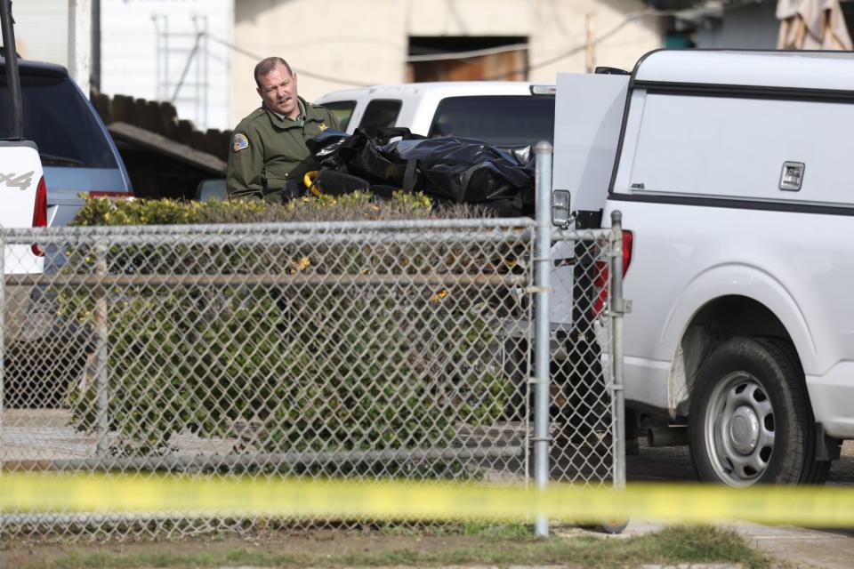 A uniformed man watches a long, horizontal black bag behind a vehicle.