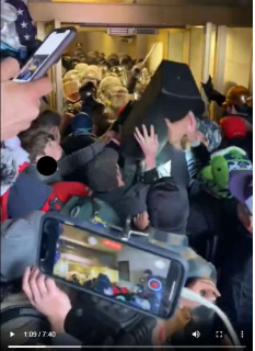 Kyle Young, seen from behind wearing a dark baseball cap, and another rioter hurl a large speaker toward a line of police defending a tunnel leading into the U.S. Capitol in a screenshot from a video taken Jan. 6, 2021. The speaker struck another member of the mob in the head, drawing blood.