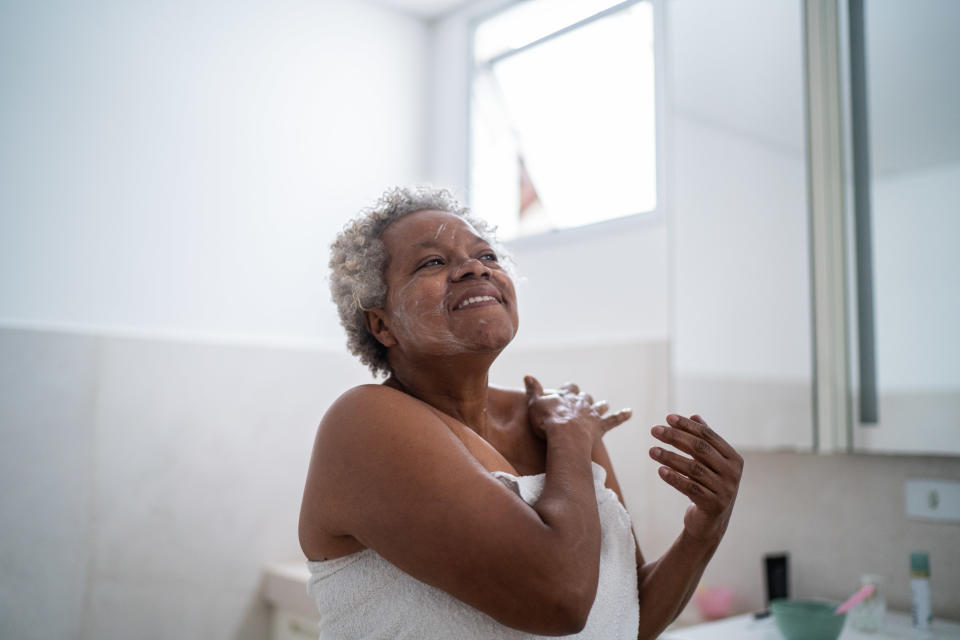 A woman with a towel wrapped around her puts moisturizer on her face in the bathroom