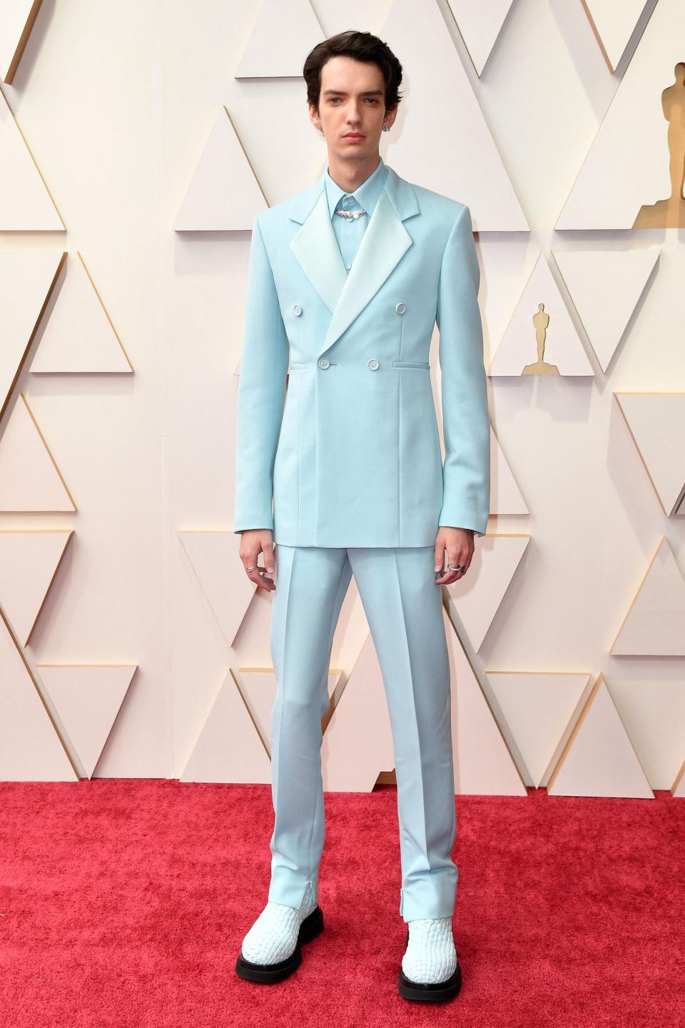 Kodi in a light blue double breast suit with a matching shirt and necklace under his collar, matching pants, and white-blue woven chunky shoes.