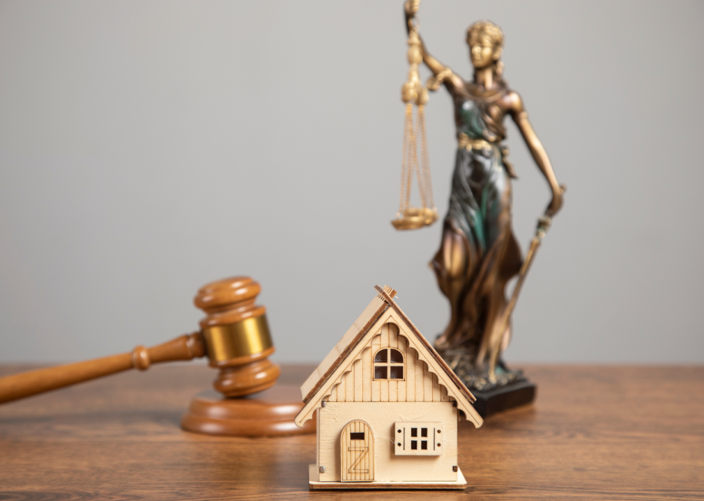 On desk sit a small wooden house, a judge's gavel, and a bronze statue of Lady Justice