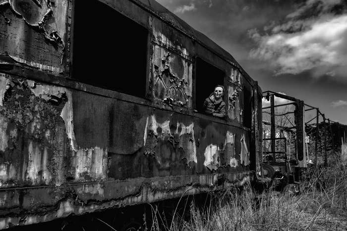 Person in a weathered train car, peering through a window in a desolate, overgrown landscape