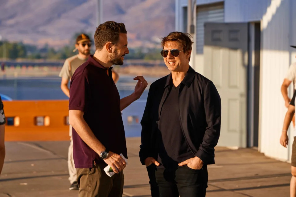 Two men, including Tom Cruise, are having a conversation outside near a building and a waterfront. Tom Cruise is wearing sunglasses and a casual jacket