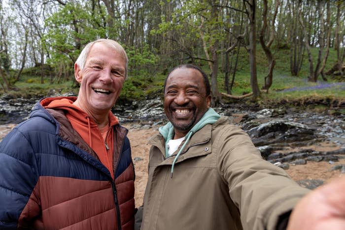 Two people smiling warmly at the camera, outdoors with trees in the background