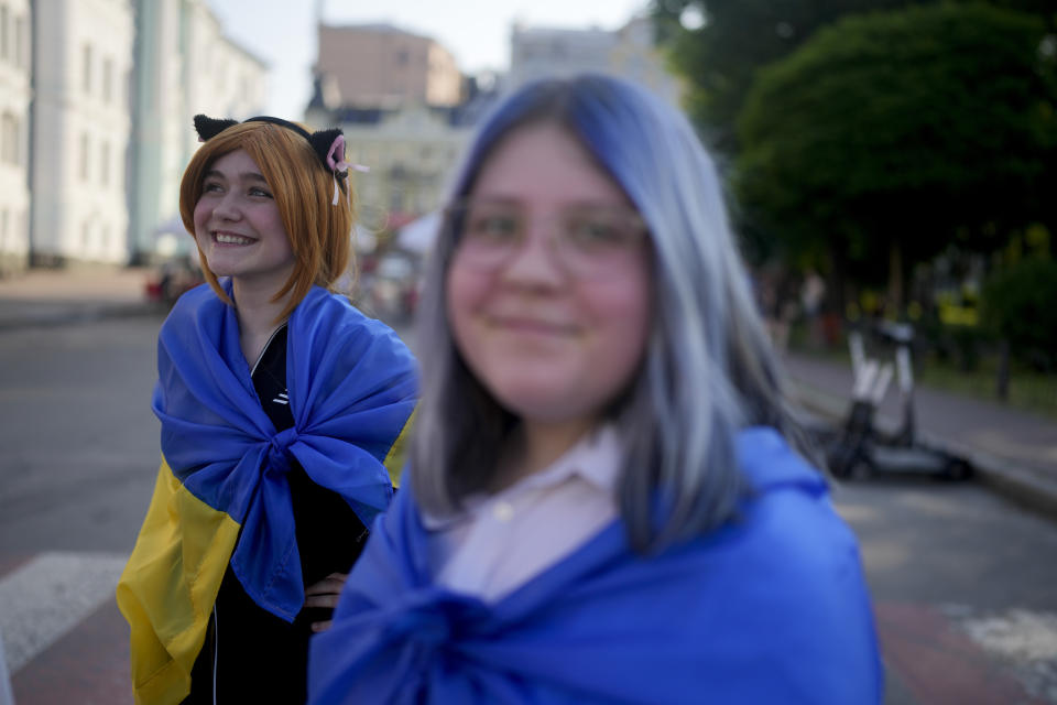Women wrapped in Ukrainian flags smile in Kyiv, Ukraine, Friday, June 10, 2022. With war raging on fronts to the east and south, the summer of 2022 is proving bitter for the Ukrainian capital, Kyiv. The sun shines but sadness and grim determination reign. (AP Photo/Natacha Pisarenko)