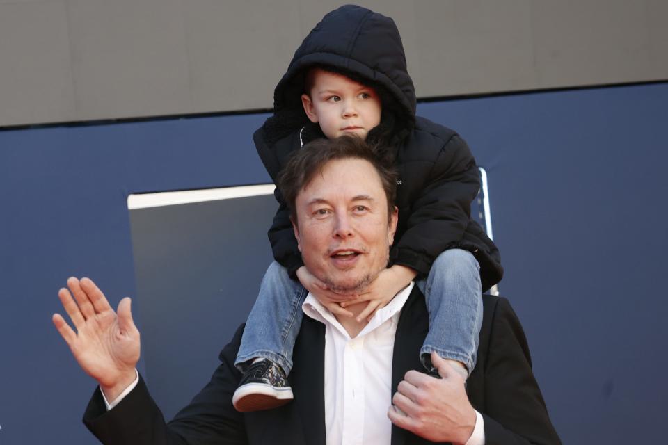 3-year-old X in a coat with a hood sitting on Musk's shoulders.