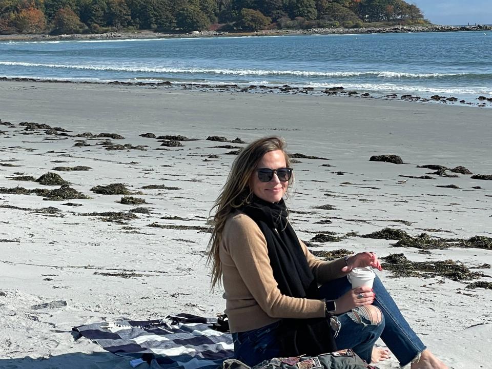 Emily, in a long-sleeve shirt, black scarf, jeans, and sunglasses, sits on a beach while holding a coffee cup.
