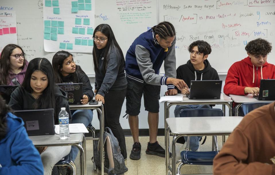 Standing between classroom desks, two peer mentors help other students during a class.