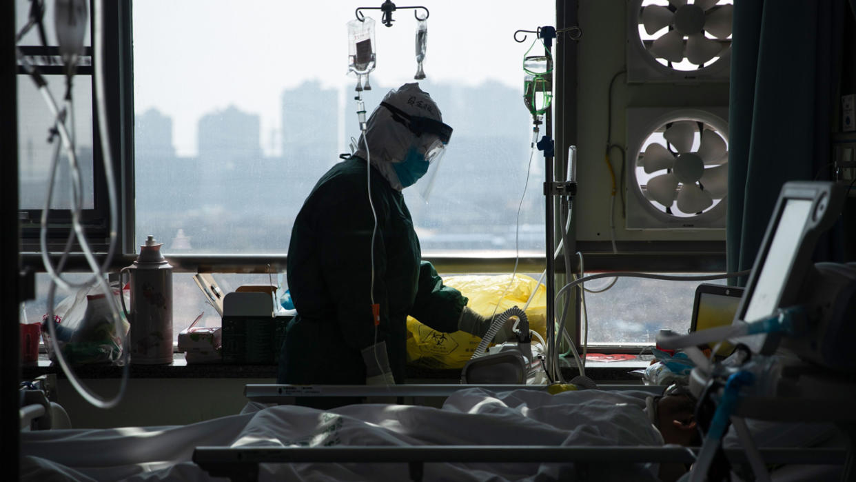 A medical staff member with hood, googles, face mask, face shield and rubber gloves tends to a person in a hospital bed.